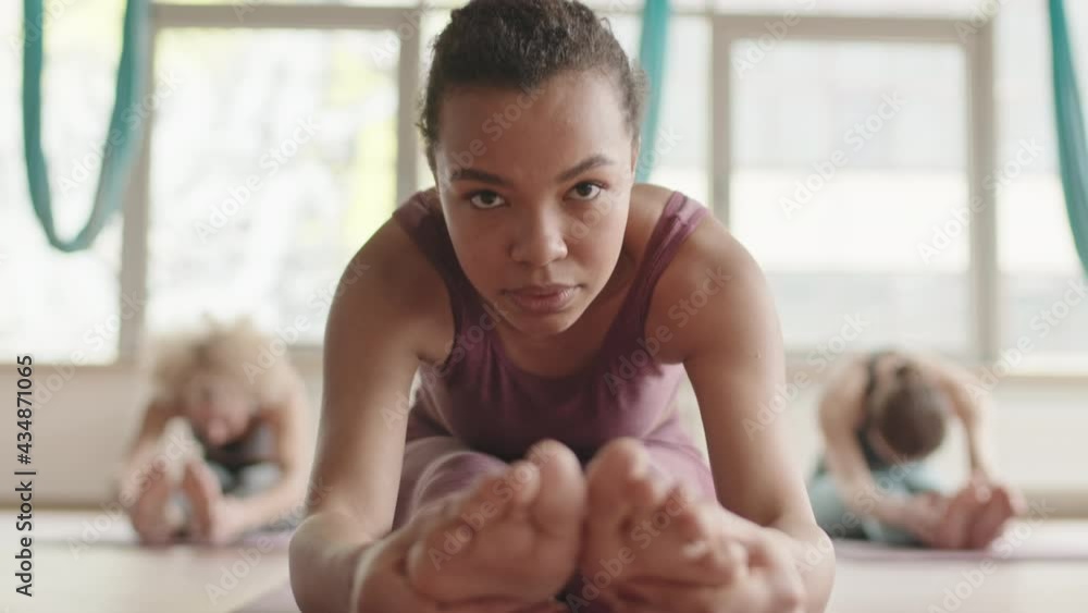 Locked-down of young Mixed-Race woman wearing sports clothing sitting with straight legs on yoga mat, folding forward and sitting back up, blurred people exercising on background