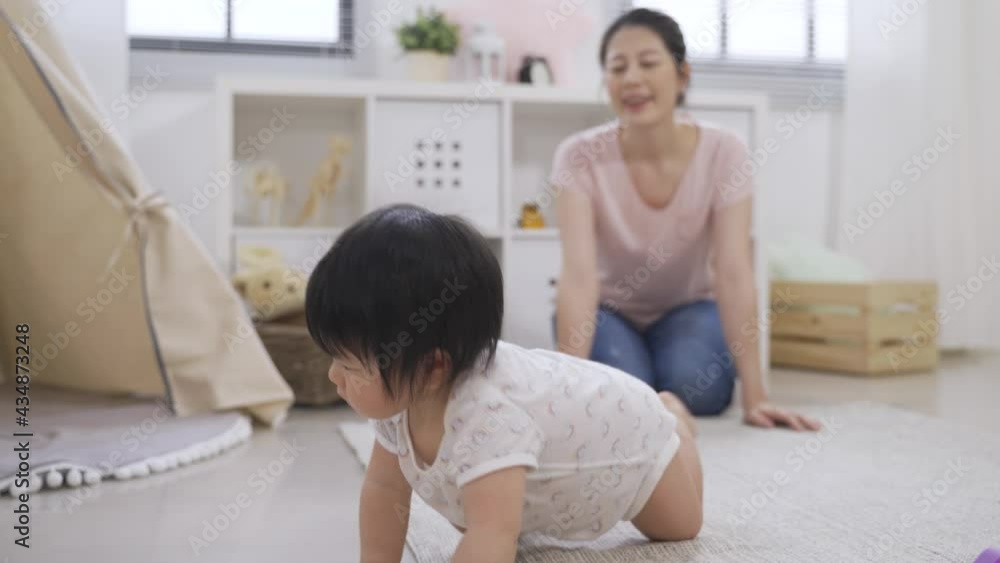 cheerful Korean mom is spending time with her young child who’s crawling slowly on the floor in a cozy bright playroom at home. cheerful Korean mom is spending time with her young child who’s crawling slowly on the floor in a cozy bright playroom at home.
