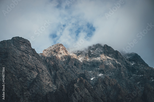 Atmospheric mountain landscape with low cloud on mountain top in sunlight. Dark rocks with snow and blue skylight in gray cloudy sky. Beautiful mountain scenery with low cloud on rocky top in sunshine