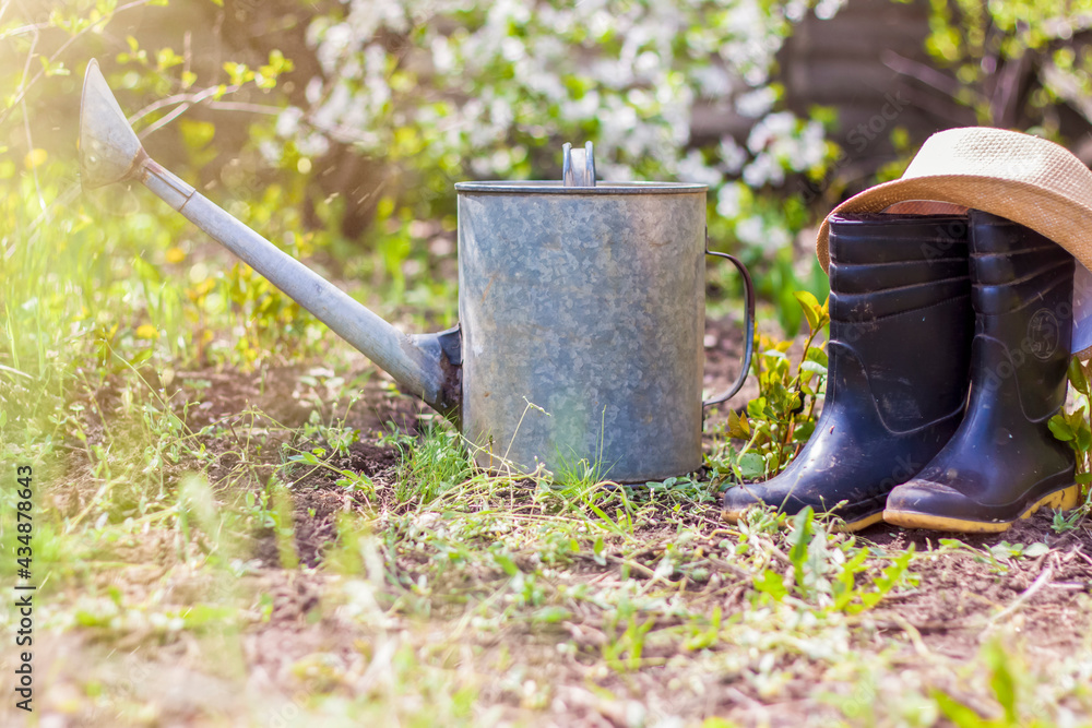 An old iron watering can, a hat, and rubber boots stand in the garden. Gardening and gardening, An old iron watering can, a hat, and rubber boots stand in the garden. Gardening and gardening,