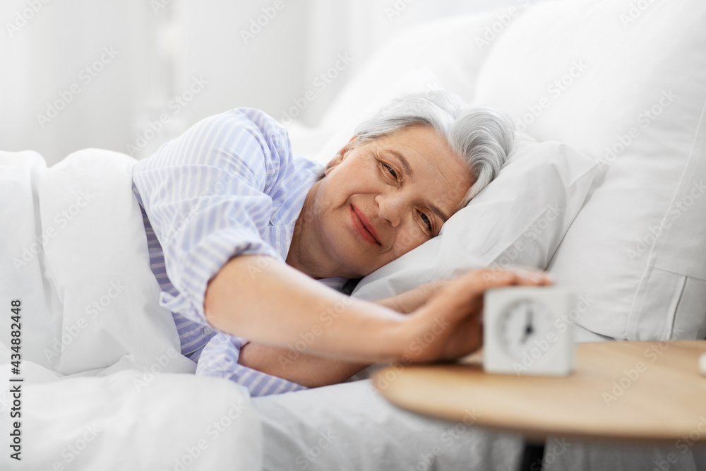 old age and people concept - happy smiling senior woman with alarm clock lying in bed at home bedroom old age and people concept - happy smiling senior woman with alarm clock lying in bed at home bedroom