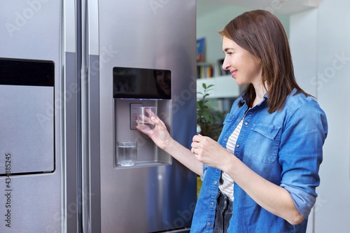 Water dispenser, woman taking cold water into glass from home refrigerator photo