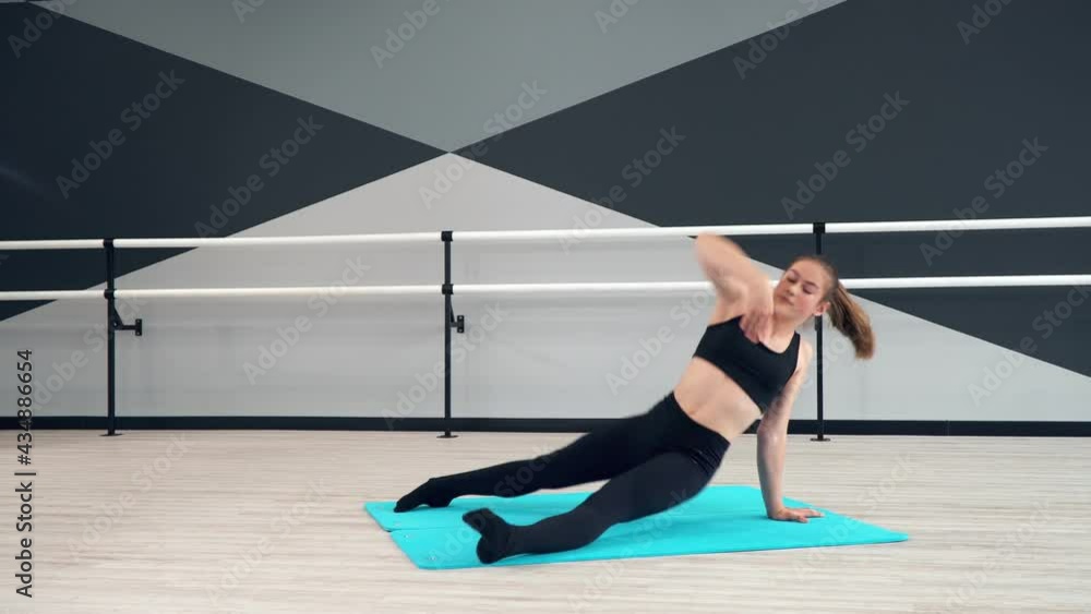 Motion of young pretty flexible girl practicing choreographic move on floor. Attractive fit female dancer stretching on mat in dance hall with handrials, wall with geometric painting on background. Motion of young pretty flexible girl practicing choreographic move on floor. Attractive fit female dancer stretching on mat in dance hall with handrials, wall with geometric painting on background.
