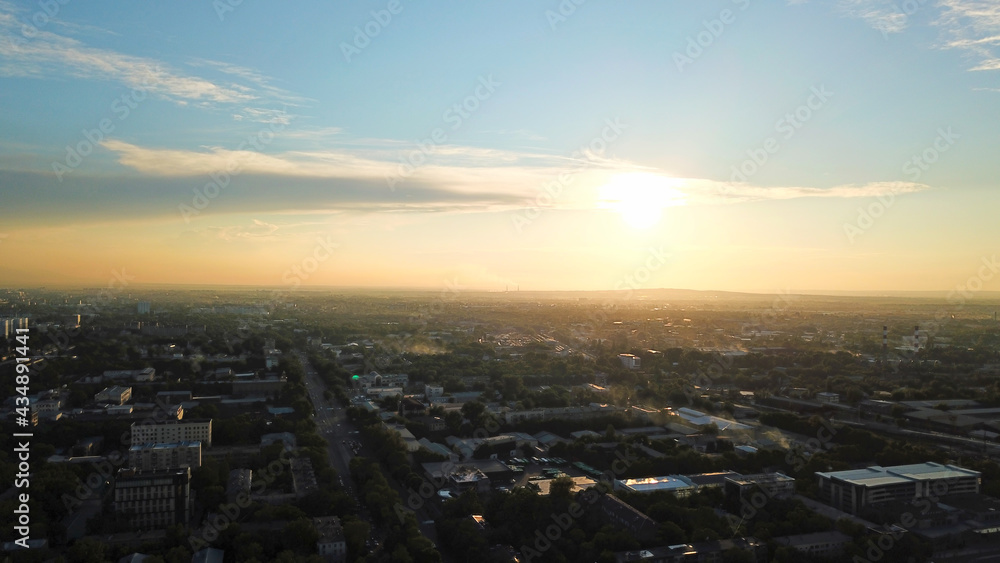 Bright color sunset over the city of Almaty. Huge clouds over the mountains and the city shimmer from bright blue to yellow and dark blue. Tall houses and green trees, cars driving on the roads.