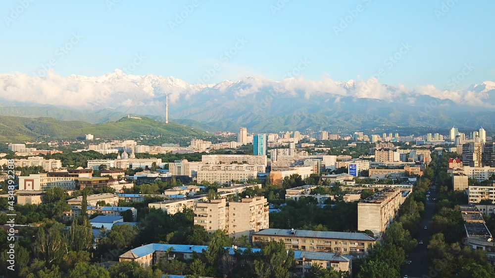 Bright color sunset over the city of Almaty. Huge clouds over the mountains and the city shimmer from bright blue to yellow and dark blue. Tall houses and green trees, cars driving on the roads.