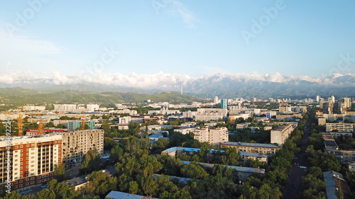 Bright color sunset over the city of Almaty. Huge clouds over the mountains and the city shimmer from bright blue to yellow and dark blue. Tall houses and green trees, cars driving on the roads.