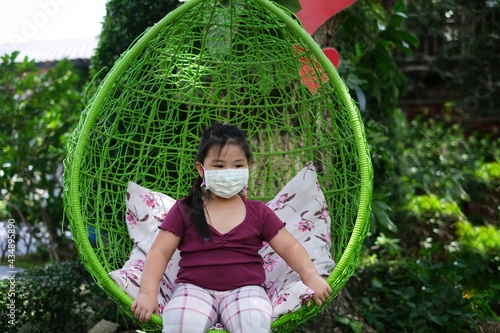 A cute young chubby Asian girl is sitting in a green round rattan swing chair with pillows in a garden, resting and relaxing.