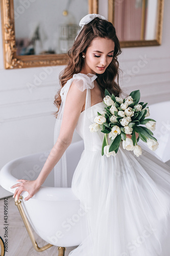 Beautiful bride with a wedding bouquet sitting on sofa indoors in white studio interior like at home. photo