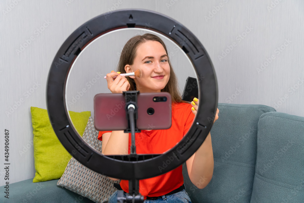 Young, cheerful, beautiful, dark-haired woman records a video about cosmetics or leads a beauty stream on a mobile phone camera with additional lighting with a ring lamp Young, cheerful, beautiful, dark-haired woman records a video about cosmetics or leads a beauty stream on a mobile phone camera with additional lighting with a ring lamp