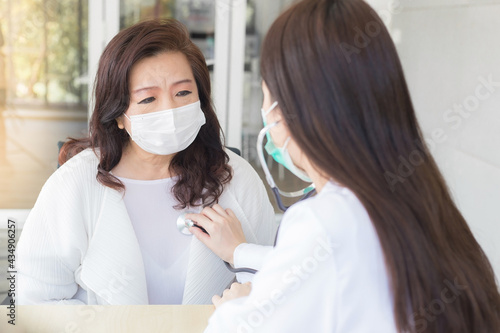 Asian beautiful female doctor uses stethoscope to check health of lungs and heart or diagnosis symptom of woman  elderly patient while both wear face mask in hospital.