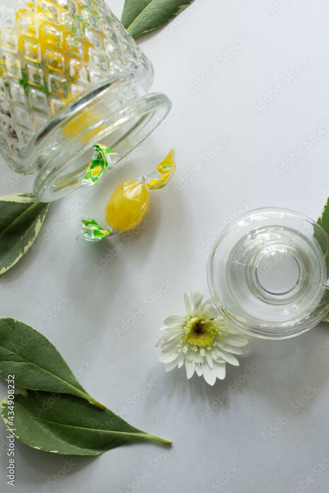 Still life with a glass vessel, white flowers, leaves and yellow sweets on a white table Still life with a glass vessel, white flowers, leaves and yellow sweets on a white table