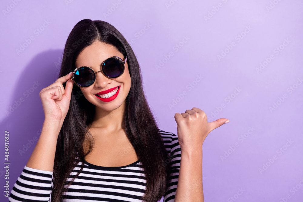 Photo of excited pretty young lady wear striped shirt arm dark eyewear pointing finger empty space isolated purple color background Photo of excited pretty young lady wear striped shirt arm dark eyewear pointing finger empty space isolated purple color background
