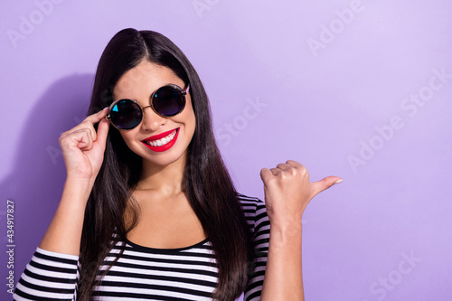 Photo of excited pretty young lady wear striped shirt arm dark eyewear pointing finger empty space isolated purple color background