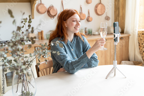 Happy online remote celebration on distance. Smiling ginger haired girl holding glass of champagne and looking at mobile phone screen. Video call chatting pandemic time. Long distance relationship  photo
