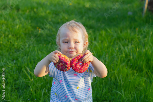 Beautiful baby girl 2 years old closes her eyes with two pink donuts in the summer in nature