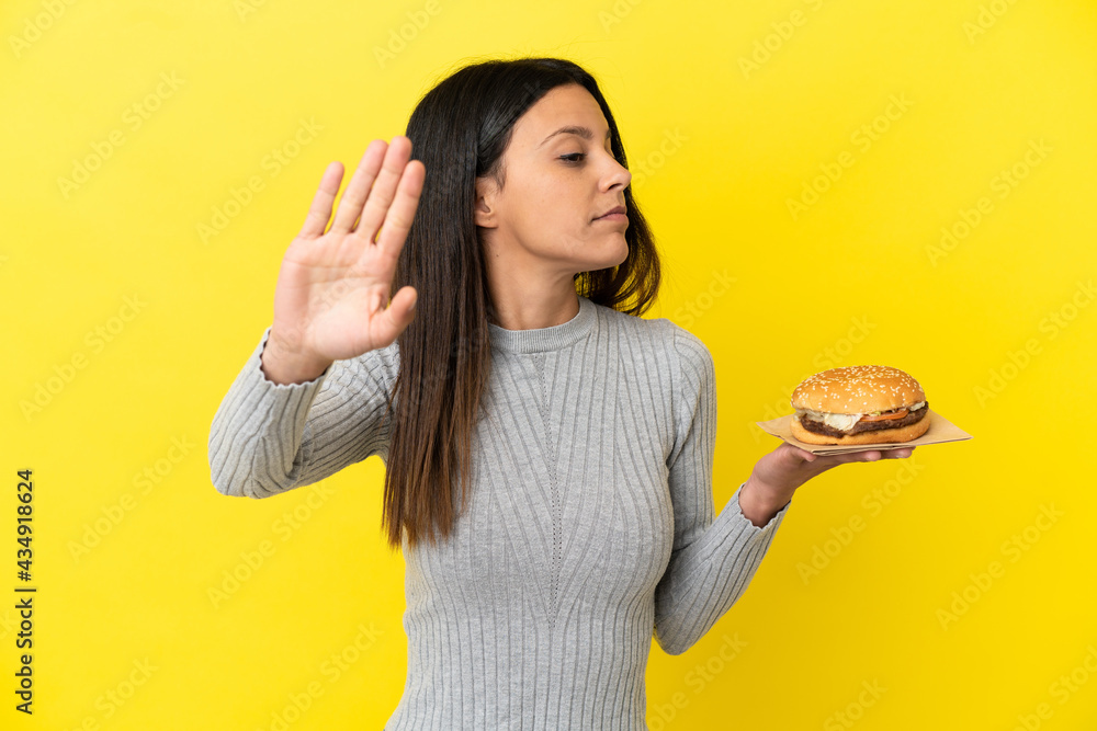 Young caucasian woman holding a burger isolated on yellow background making stop gesture and disappointed Young caucasian woman holding a burger isolated on yellow background making stop gesture and disappointed