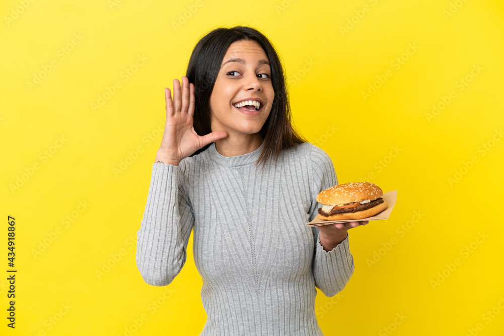 Young caucasian woman holding a burger isolated on yellow background listening to something by putting hand on the ear Young caucasian woman holding a burger isolated on yellow background listening to something by putting hand on the ear