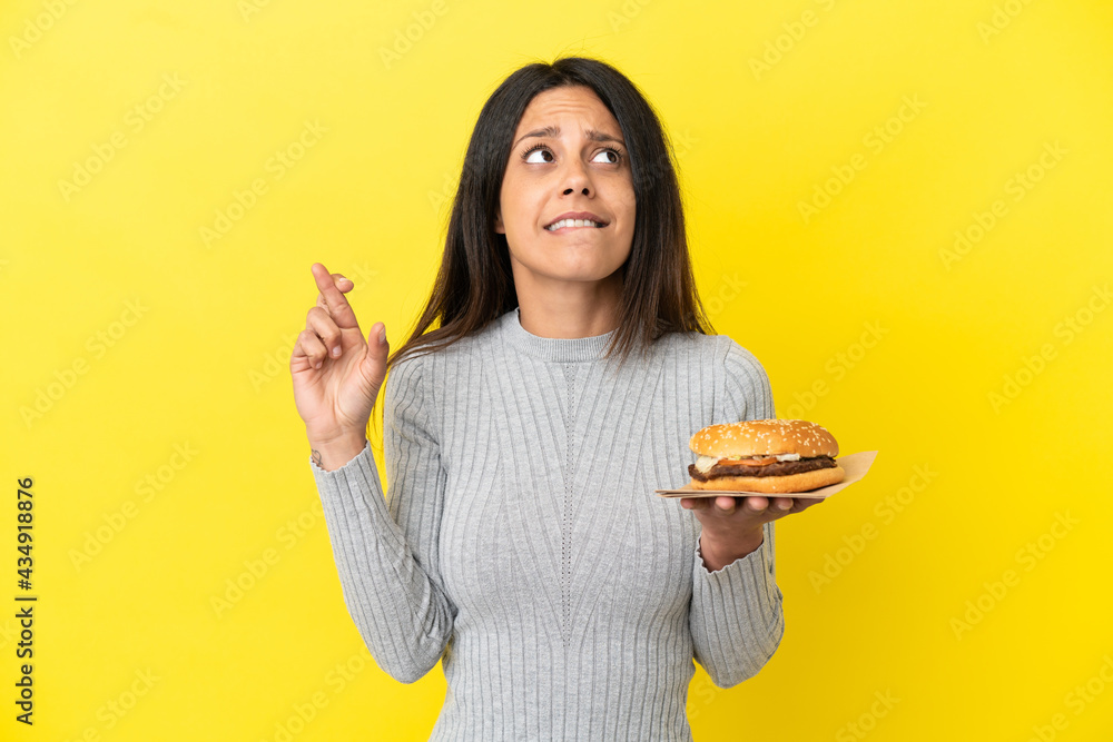 Young caucasian woman holding a burger isolated on yellow background with fingers crossing and wishing the best Young caucasian woman holding a burger isolated on yellow background with fingers crossing and wishing the best