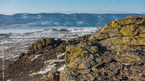 The granite rock is crossed by cracks. Yellow lichens grow on the stones. Snow on the ground. In the distance there is a mountain range, a frozen lake. Sunny day. Baikal