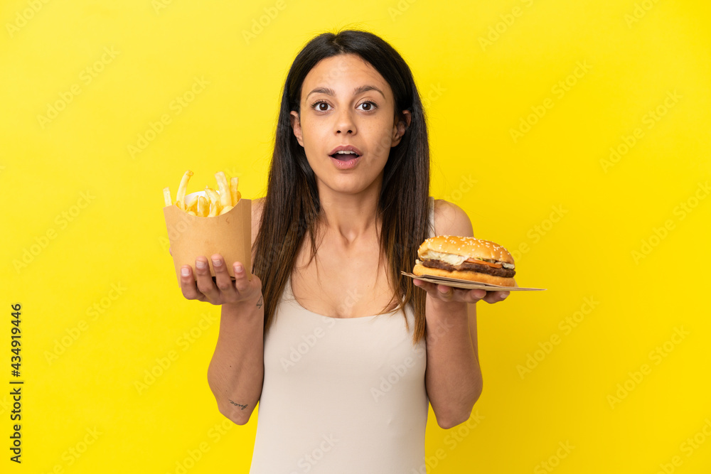 Young caucasian woman holding burger and chips isolated on yellow background Young caucasian woman holding burger and chips isolated on yellow background