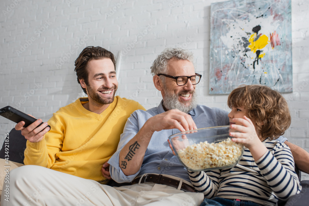 Smiling man with remote controller sitting near father and son with popcorn on couch Smiling man with remote controller sitting near father and son with popcorn on couch