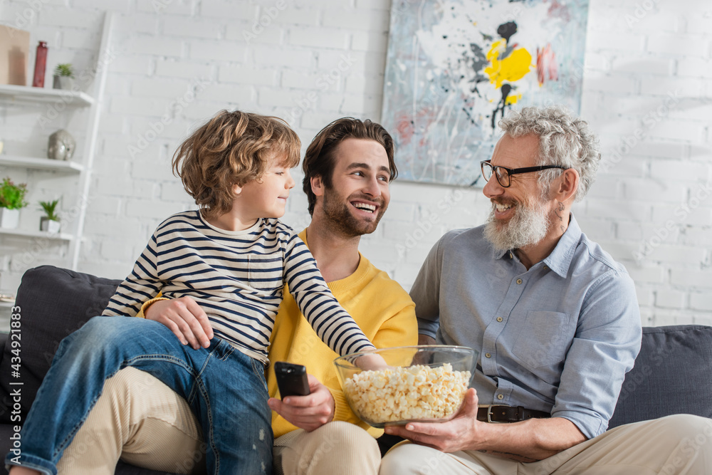 © LIGHTFIELD STUDIOS - Smiling father with remote controller and grandfather with popcorn near child on couch