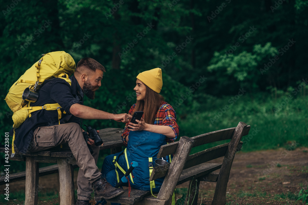 A couple of hikers sitting on a bench in the forest and making a break while using a smartphone A couple of hikers sitting on a bench in the forest and making a break while using a smartphone