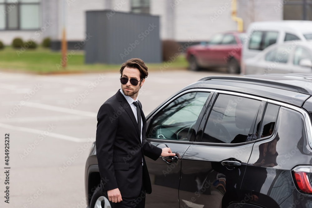 bodyguard in suit and sunglasses with security earpiece opening door of modern auto