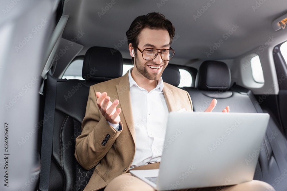 smiling businessman in glasses and wireless earphones gesturing during video call in car