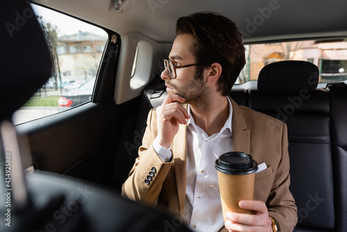 pensive man in suit and glasses holding paper cup while looking at car window