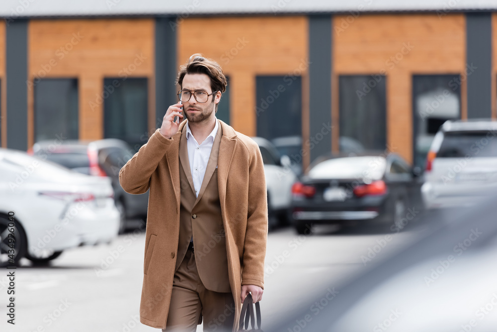 confident man in glasses and beige coat talking on mobile phone in outdoor parking confident man in glasses and beige coat talking on mobile phone in outdoor parking