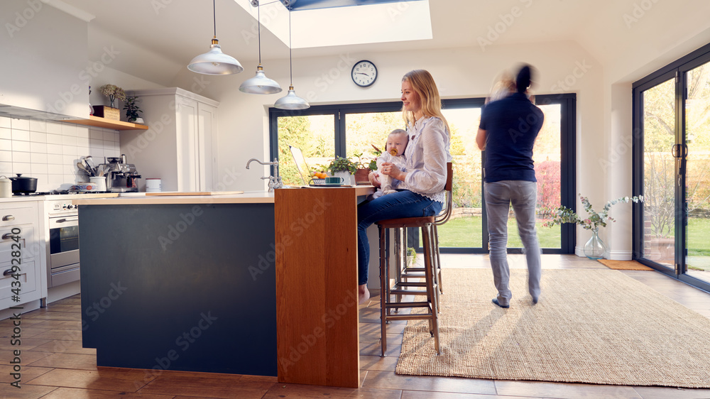Busy Family Kitchen With Mother Working On Laptop And Holding Baby Whilst Father Cuddles Daughter Busy Family Kitchen With Mother Working On Laptop And Holding Baby Whilst Father Cuddles Daughter