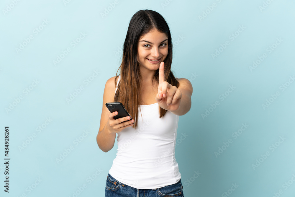 Young caucasian woman using mobile phone isolated on blue background showing and lifting a finger