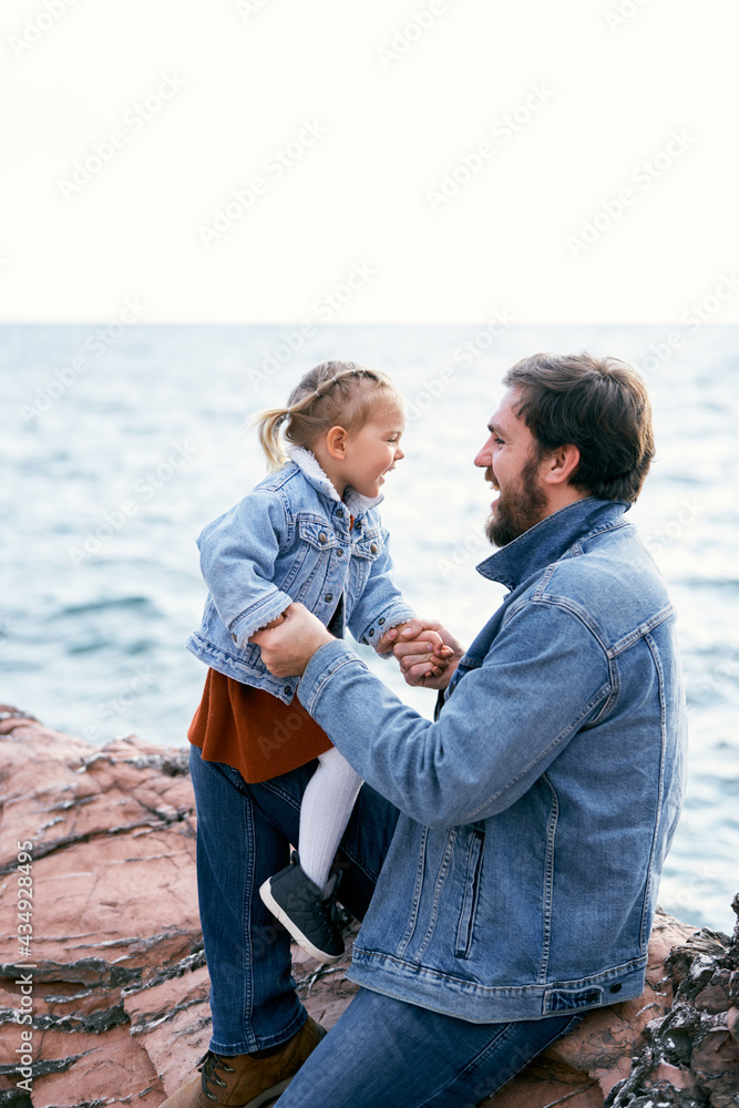 Laughing dad and little daughter sit on stones face to face near the sea, holding hands