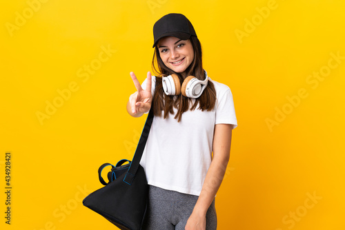 Young sport woman with sport bag isolated on yellow background smiling and showing victory sign