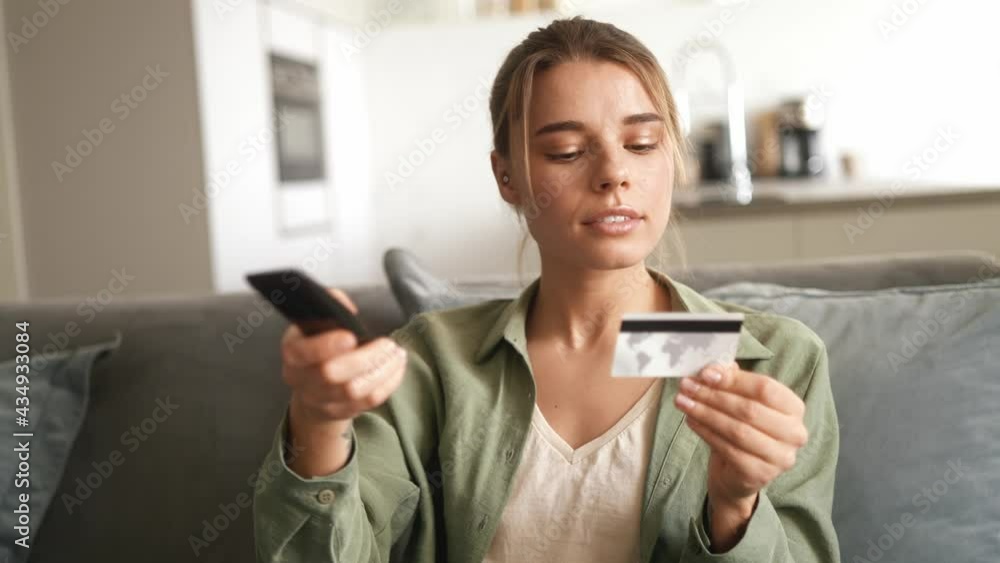The smiling woman entering a bank card number on the TV and using the remote control while sitting on the couch in the room The smiling woman entering a bank card number on the TV and using the remote control while sitting on the couch in the room