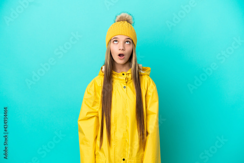 Little girl wearing a rainproof coat over isolated blue background looking up and with surprised expression © luismolinero