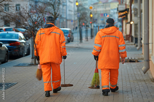 Street sweaper clean road and pedestrian zone in the city. Municipal workers sweep city street, janitors with broomstick and scoop for garbage. Men in orange uniform collecting garbage from sidewalk