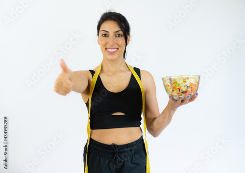 Profile portrait positive healthy woman in white sportswear holding big bowl with fresh vegetable salad and measure tape. Healthy nutrition.