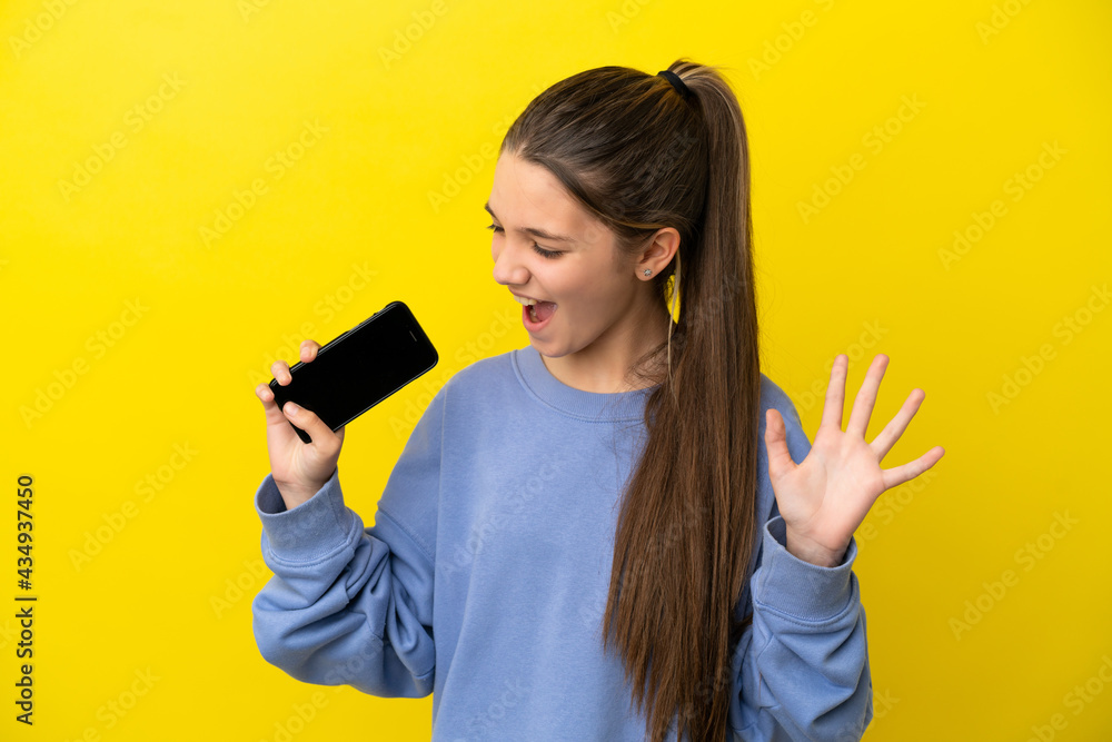 Little girl over isolated yellow background using mobile phone and singing Little girl over isolated yellow background using mobile phone and singing
