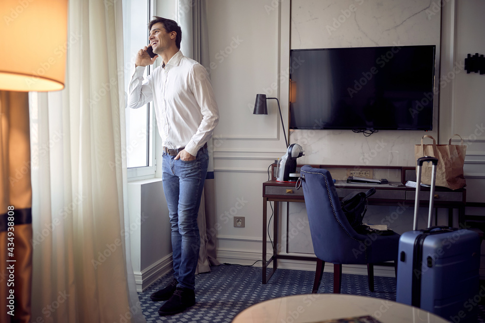 handsome young caucasian man talking on cell phone in a hotel room, standing by the window, smiling