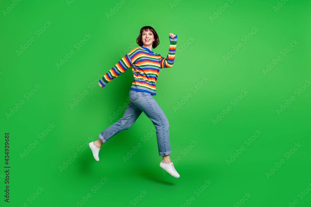 Full length photo of funky sweet young woman wear striped sweater smiling jumping high running isolated green color background