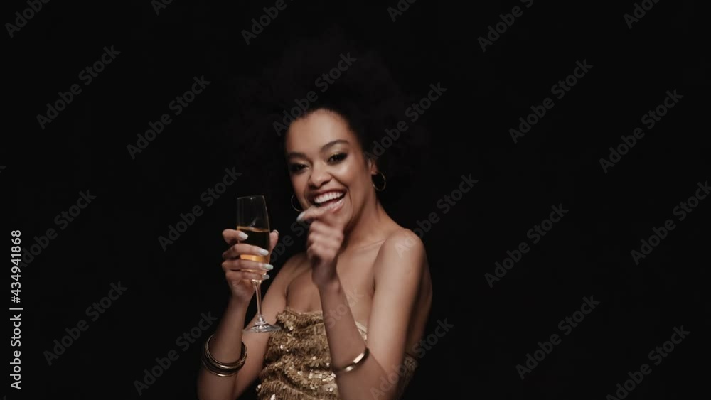 The smiling woman dancing with a glass in her hands and showing peace gesture in a black studio The smiling woman dancing with a glass in her hands and showing peace gesture in a black studio