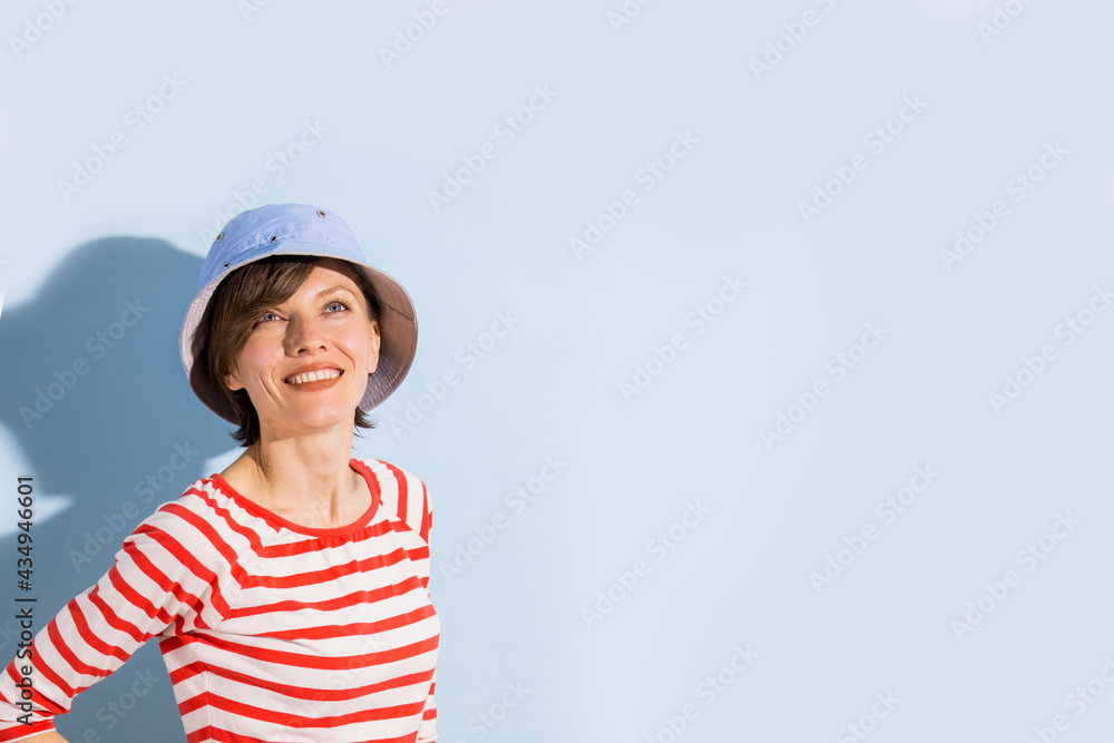 Happy cheerful young woman with short hair, natural makeup over light blue background. Indoor portrait of beautiful brunette young woman wearing blue hat and bright striped clothing Happy cheerful young woman with short hair, natural makeup over light blue background. Indoor portrait of beautiful brunette young woman wearing blue hat and bright striped clothing