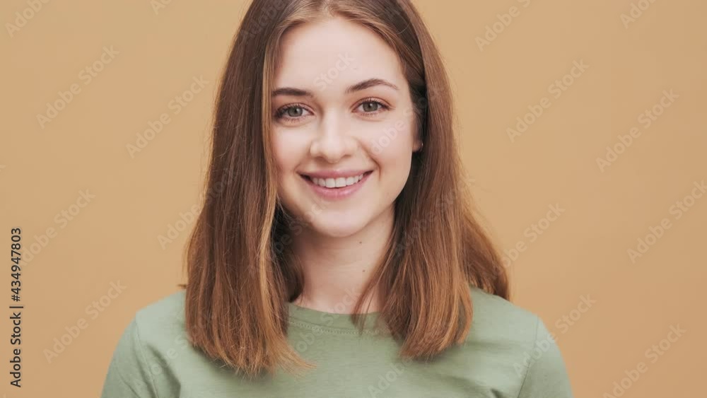 A beautiful young woman is opening her eyes and smiling to the camera standing isolated over a beige wall in the studio A beautiful young woman is opening her eyes and smiling to the camera standing isolated over a beige wall in the studio