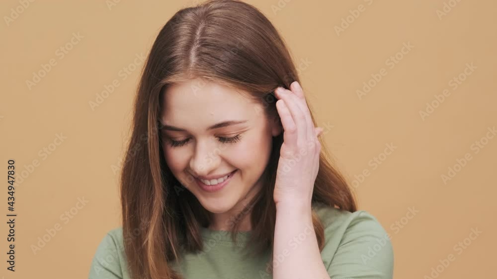 A charming young woman is smiling to the camera standing isolated over a beige wall in the studio A charming young woman is smiling to the camera standing isolated over a beige wall in the studio