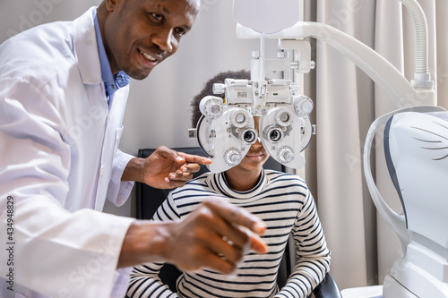 African young woman girl doing eye test checking examination with male man optometrist using phoropter in clinic or optical shop. Eyecare concept. photo