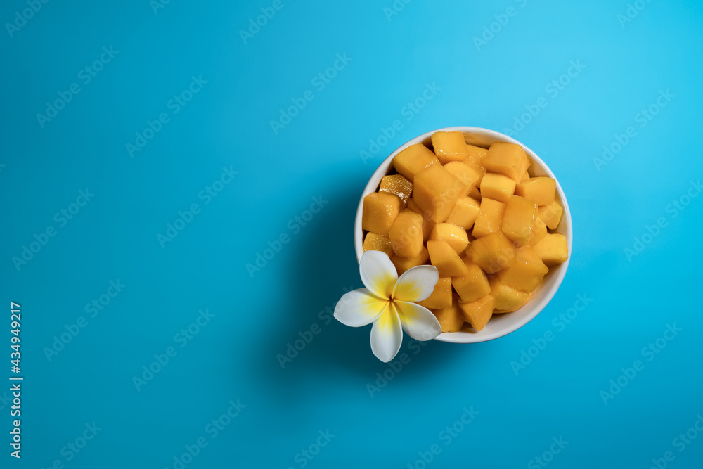 Tropical fruit concept. Yellow mango cube slices in white bowl on bright blue background with white flower. Flat lay, top view, copy space Tropical fruit concept. Yellow mango cube slices in white bowl on bright blue background with white flower. Flat lay, top view, copy space