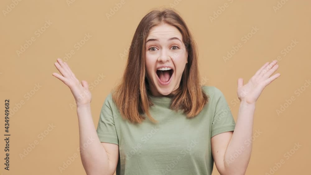 A smiling young woman is looking to the camera while raising her hands standing isolated over a beige wall in the studio A smiling young woman is looking to the camera while raising her hands standing isolated over a beige wall in the studio