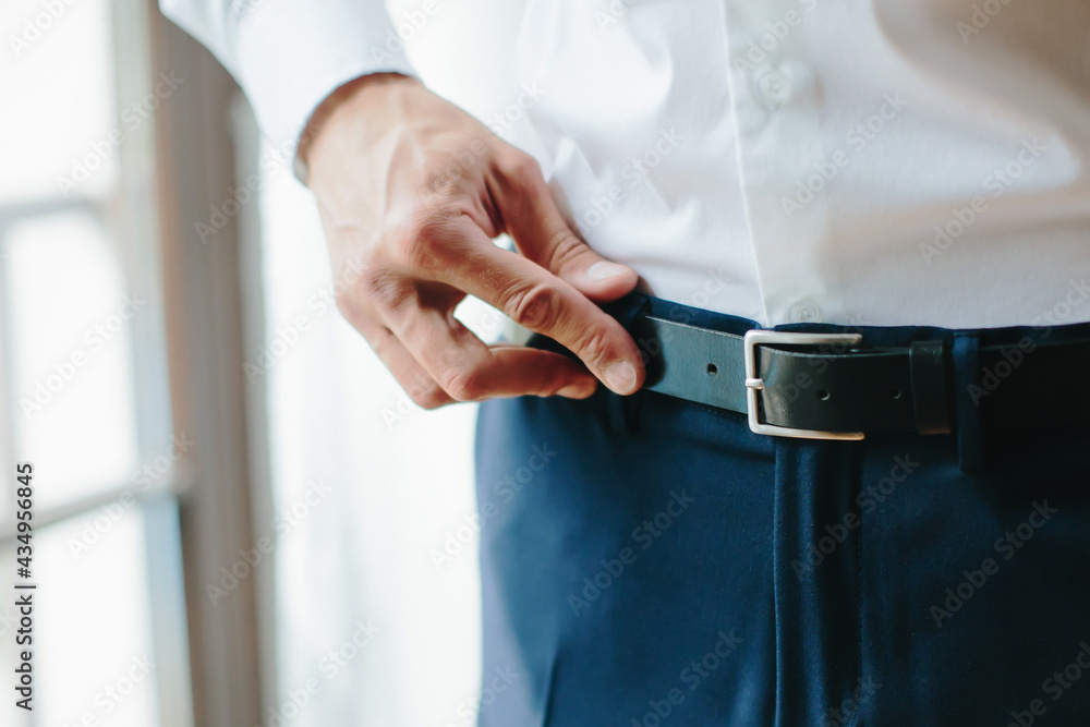 Young man fastens black belt on his trousers, male hands close up. Morning of groom, preparations for wedding. Selective focus. Young man fastens black belt on his trousers, male hands close up. Morning of groom, preparations for wedding. Selective focus.